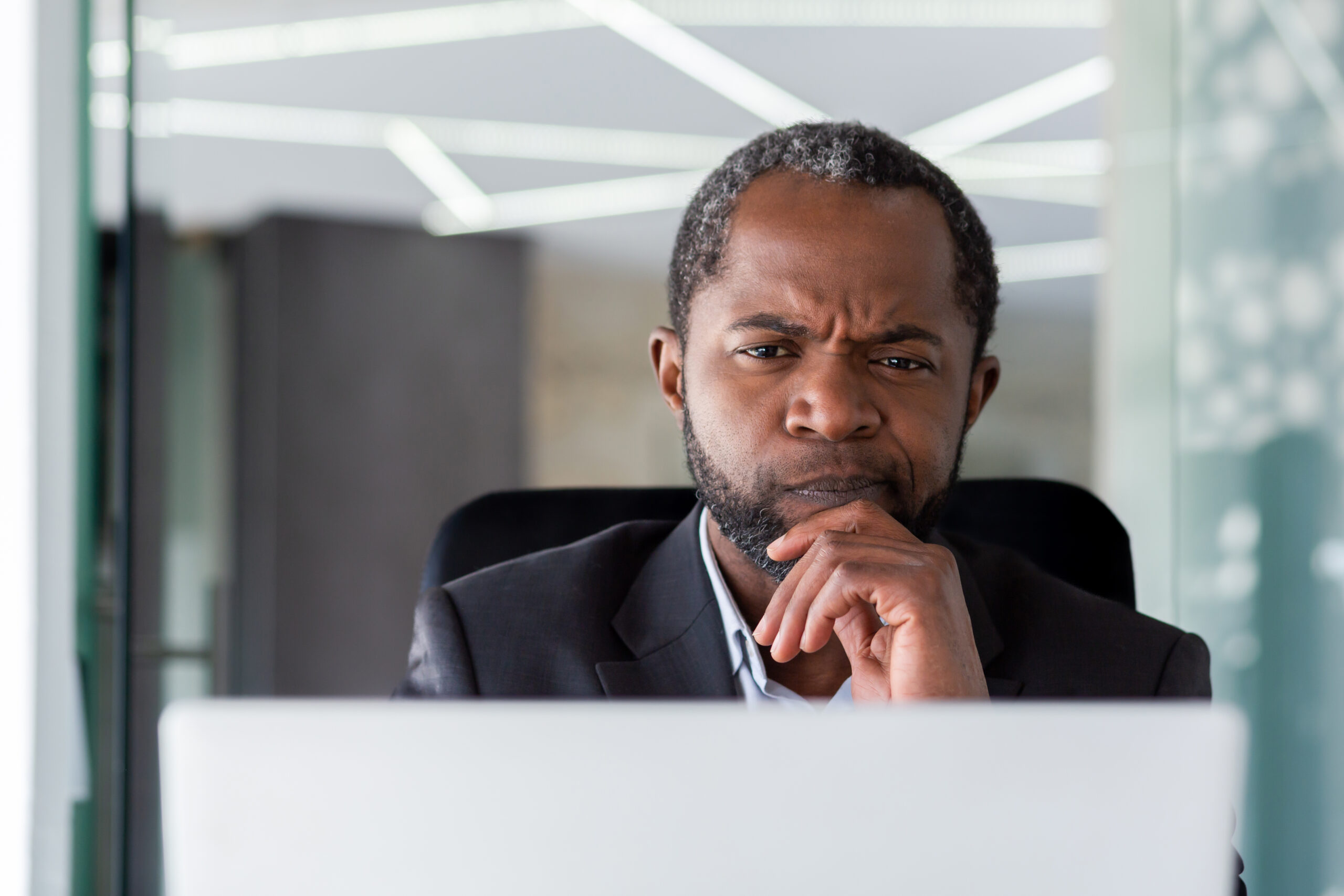 Business professional in a dark suit thoughtfully reviewing information on a laptop in a modern office, reflecting critical thinking and independent judgment.